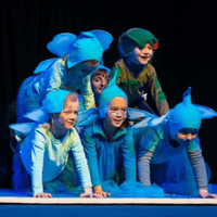 Sechs Kinder stellen sich im Vierfüßlerstand gemeinsam zu einer Pyramide auf. Fünf Kinder tragen ein blaues Feenkostüm, ein Kind ist als Pflanze verkleidet.