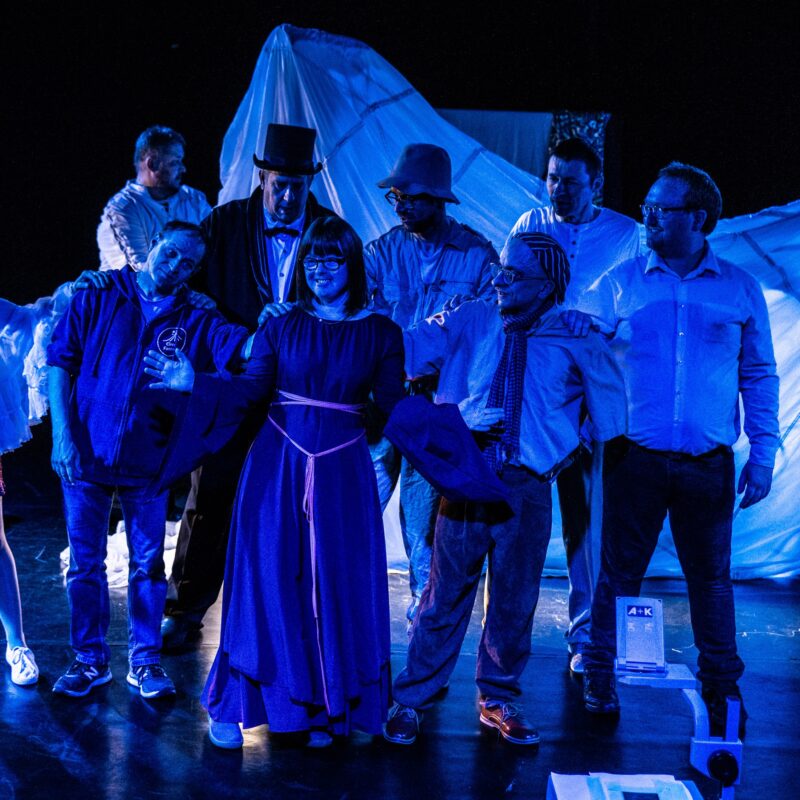 Eine Gruppe von Schauspielenden steht in Kleider und Hemden auf der Bühne, auf sie scheint ein blaues Licht