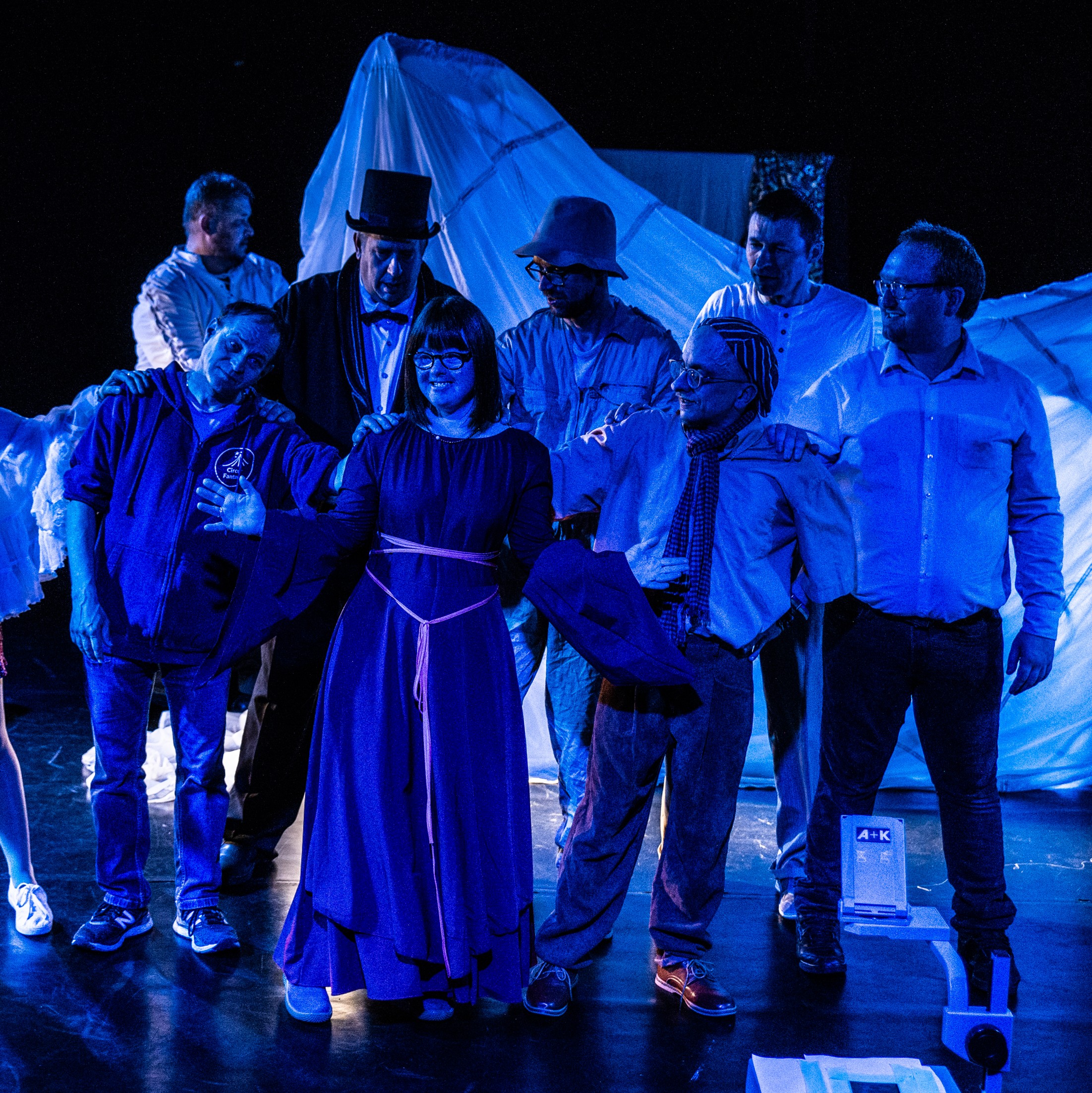 Eine Gruppe von Schauspielenden steht in Kleider und Hemden auf der Bühne, auf sie scheint ein blaues Licht