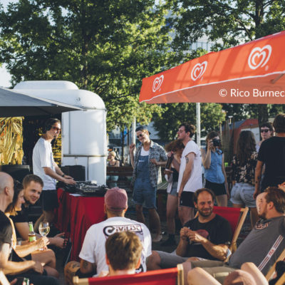 Verschiedene Menschen tanzen und sitzen unter freiem Himmel während ein DJ spielt.