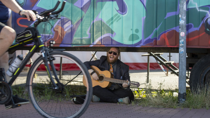 Ein Mann sitzt im Schneidersitz vor einem Circuswagen und spielt Gitarre.