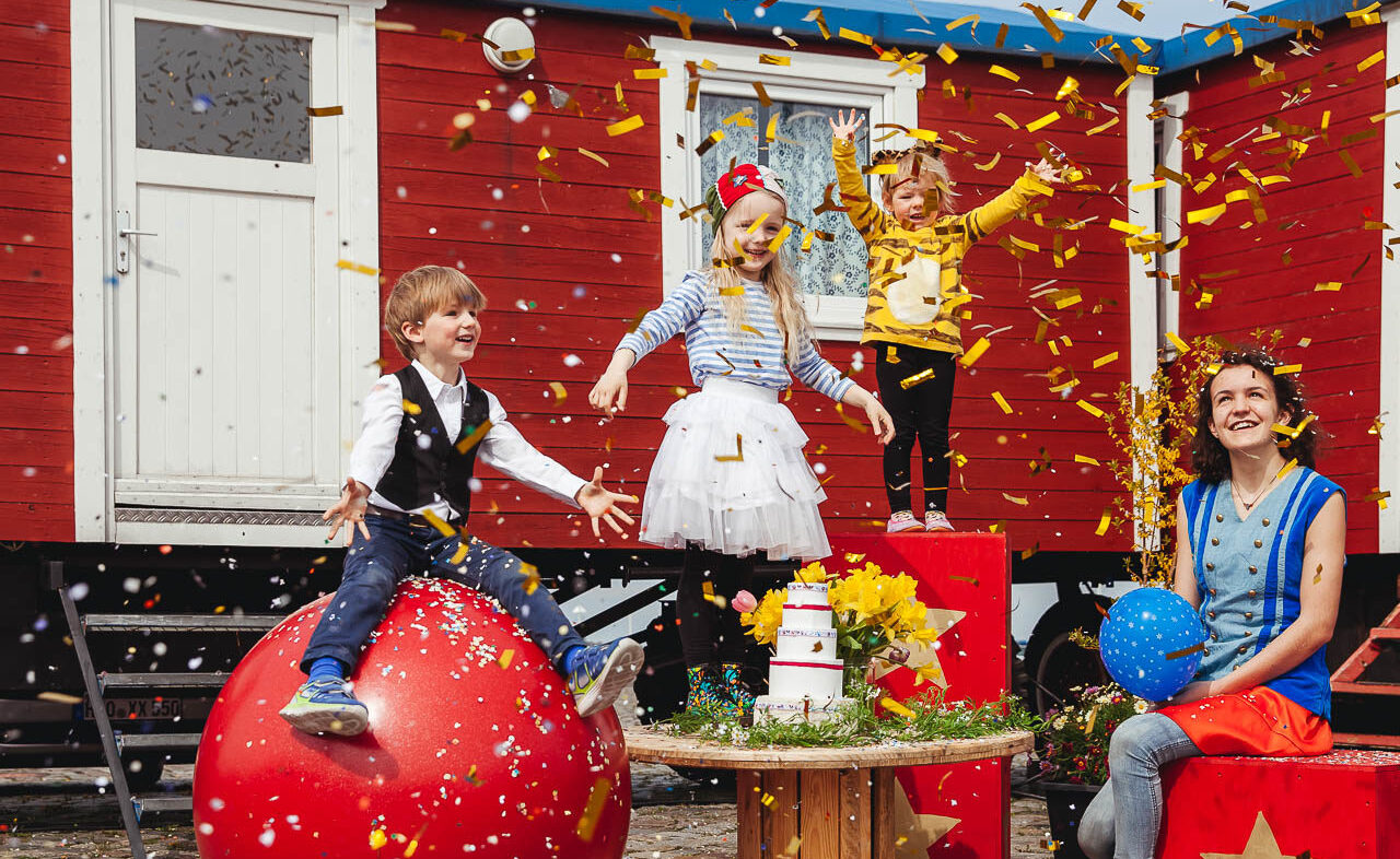 Drei Kinder und eine Frau freuen sich und schauen in die Luft. Dort fliegt Konfetti durch die Gegend. Sie sitzen auf Circusrequisiten.