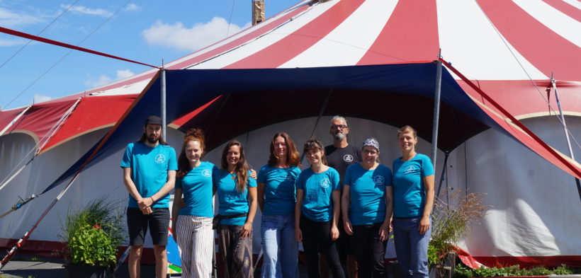 Das Team des Circus Fantasia steht nebeneinander vor dem Circuszelt und schaut lächelnd in die Kamera.