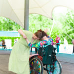 Eine Frau in einem grünen Kleid umarmt in einer Tanzchoreographie eine andere Frau in einem Rollstuhl.