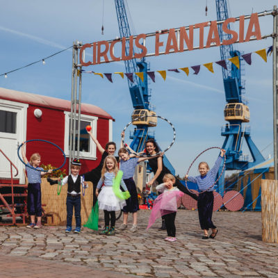 Mehrere Kinder und eine Erwachsene stehen mit Hula Hoop Reifen vor dem Eingang des Circus Fantasia Platzes.
