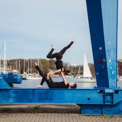 Eine Frau liegt mit ausgestrecktem Bein auf dem Rücken und stützt eine andere Person an den Schultern, die mit ausgestreckten Beinen einen Handstand über ihr macht. Im Hintergrund ist die Warnow.