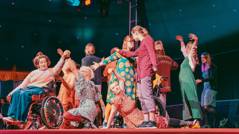 Eine Gruppe von verschiedenen Personen tanzt gemeinsam auf der Bühne. Die Gruppe berührt sich mit verschiedenen Körperteilen und ist einander zugewandt.