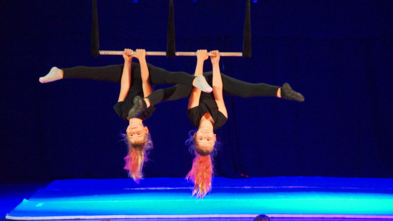 Zwei Mädchen hängen kopfüber an einem Doppel-Trapez, strecken ihre Beine aus und schauen zur Kamera.