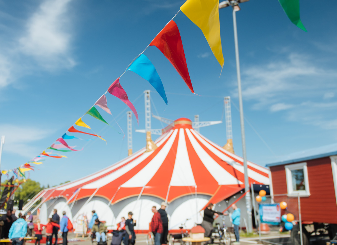 Eine bunte Wimpelkette hängt unter freiem Himmel bei Sonnenschein auf dem Circus Fantasia Platz. Im Hintergrund stehen Menschen und das große Circuszelt.