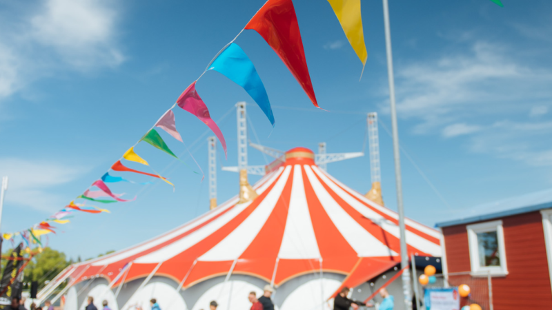 Eine bunte Wimpelkette hängt unter freiem Himmel bei Sonnenschein auf dem Circus Fantasia Platz. Im Hintergrund stehen Menschen und das große Circuszelt.