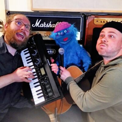 Zwei Männer mit einem Keyboard und einer Gitarre in der Hand, die lustige Grimassen ziehen & eine blaue, wuschelige Handpuppe, die freudig den Mund aufreißt
