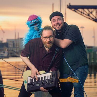 Zwei Männer mit einem Keyboard und einer Gitarre in der Hand, die lustige Grimassen ziehen & eine blaue, wuschelige Handpuppe, die freudig den Mund aufreißt, im Hintergrund ist der Rostocker Stadthafen zu sehen