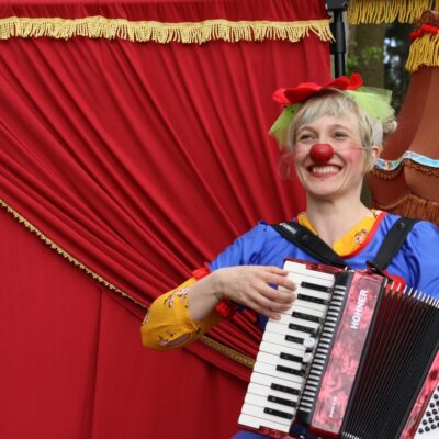 Eine Frau im Clownskostüm mit einem Akkordeon in der Hand steht vor einem roten Vorhang