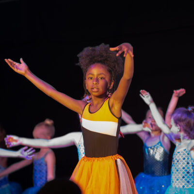 Ein Mädchen im gelben Outfit tanzt, im Hintergrund stehen weitere Mädchen, die ebenfalls tanzen