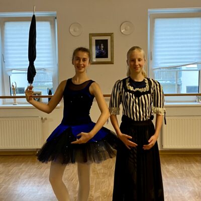 Eine Mädchen im Tutu hält einen Regenschirm, daneben steht ein weiteres Mädchen in einem langen, schwarzem Kleid