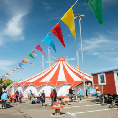 Eine Fotografie vom Circusplatz des Fantasia unter blauem Himmel, über den Platz ist eine bunte Wimpelkette gehängt und im Hintergrund ragt das Circuszelt zwischen den Circuswagen hervor