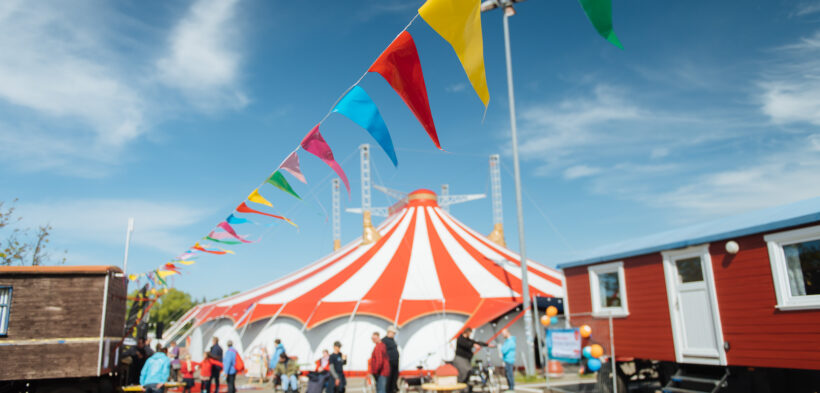 Eine Fotografie vom Circusplatz des Fantasia unter blauem Himmel, über den Platz ist eine bunte Wimpelkette gehängt und im Hintergrund ragt das Circuszelt zwischen den Circuswagen hervor