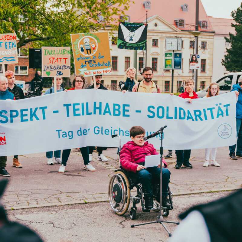Eine Person im Rollstuhl hält eine Rede, dahinter ist ein Transpi mit der Aufschrift "Respekt - Teilhabe - Solidarität" zu sehen