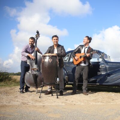 Drei Männer stehen vor einem Auto unter blauem Himmel, sie spielen dabei die Instrumente Kontrabass, Conga und Gitarre