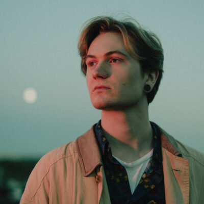 Eine Portrait-Fotografie von Joris Rose, unter freiem Himmel mit einer beigen Jacke an, den Blick in die ferne gerichtet