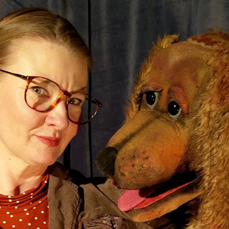 Eine Frau mit Brille schaut in die Kamera - neben ihr ein brauner, Kuscheltierhund