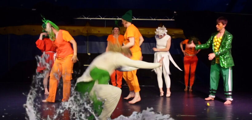 Verschiedene Menschen stehen in bunten Outfits auf der Bühne - eine Person springt in eine Pfütze