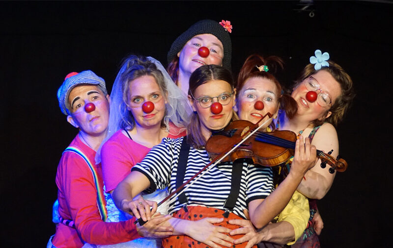 verschiedene Menschen mit Clownsnasen stehen beieinander und schauen in Richtung Kamera, ein Clown spielt eine Geige