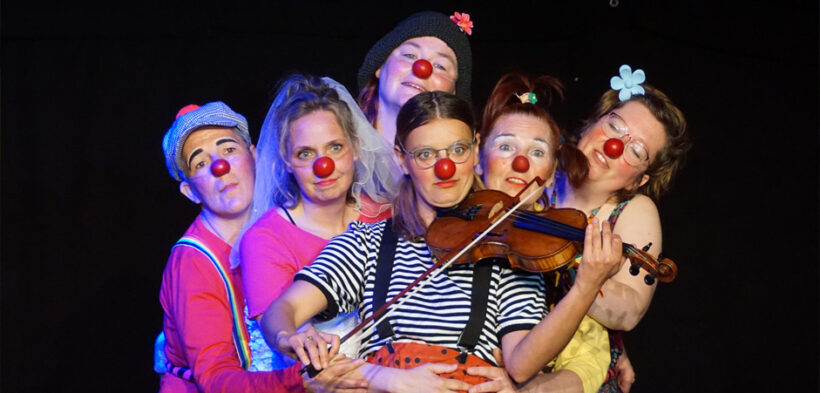 verschiedene Menschen mit Clownsnasen stehen beieinander und schauen in Richtung Kamera, ein Clown spielt eine Geige