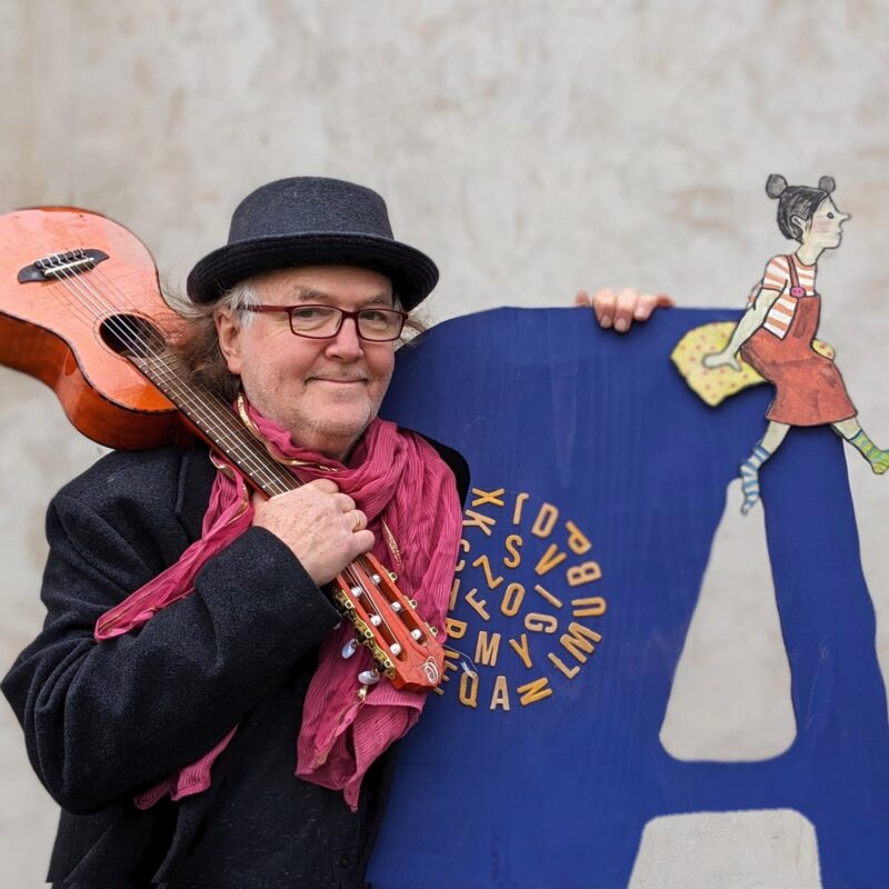 Eine Fotografie von Gerhard Schöne mit Gitarre über der Schulter und einem Lächeln, stehend vor dem Buchstaben "A"