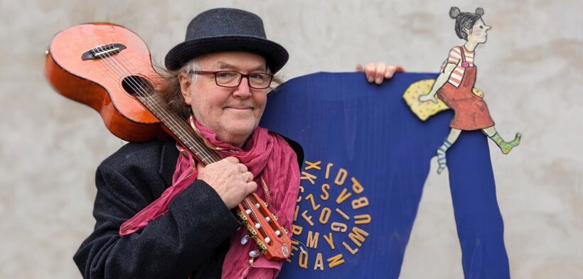 Eine Fotografie von Gerhard Schöne mit Gitarre über der Schulter und einem Lächeln, stehend vor dem Buchstaben "A"