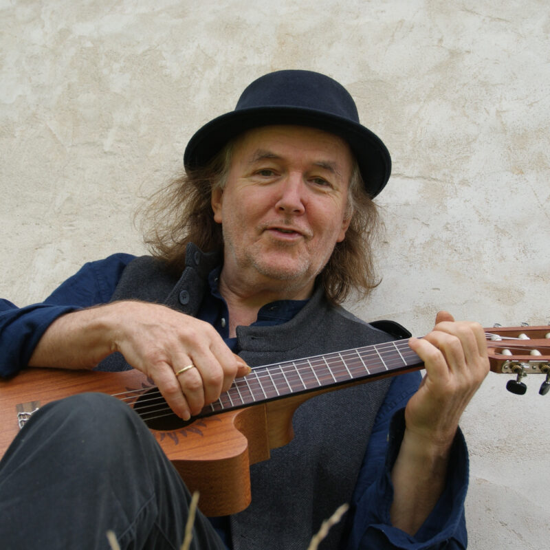 Eine Fotografie von Gerhard Schöne mit Gitarre in der Hand, schulterlangem Haar und verschmitztem Lächeln