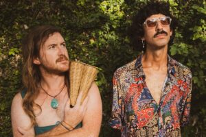 Zwei Männer mit langem Haar und Schnauzbart schauen verträumt nach oben, ihr Kleidungsstil erinnert an den Hippie-Look