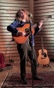 Ein Mann mit Gitarre, wirft sein langes, lockiges Haar umher und singt dabei in ein Mikrofon