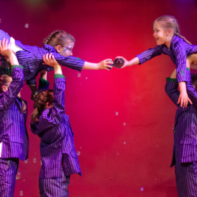 Kinder in lila Circus-Kleidung stehen in einer Reihe, stämmen zwei Mädchen in die Höhe und lassen diese eine Glaskugel an die jeweils andere übergeben