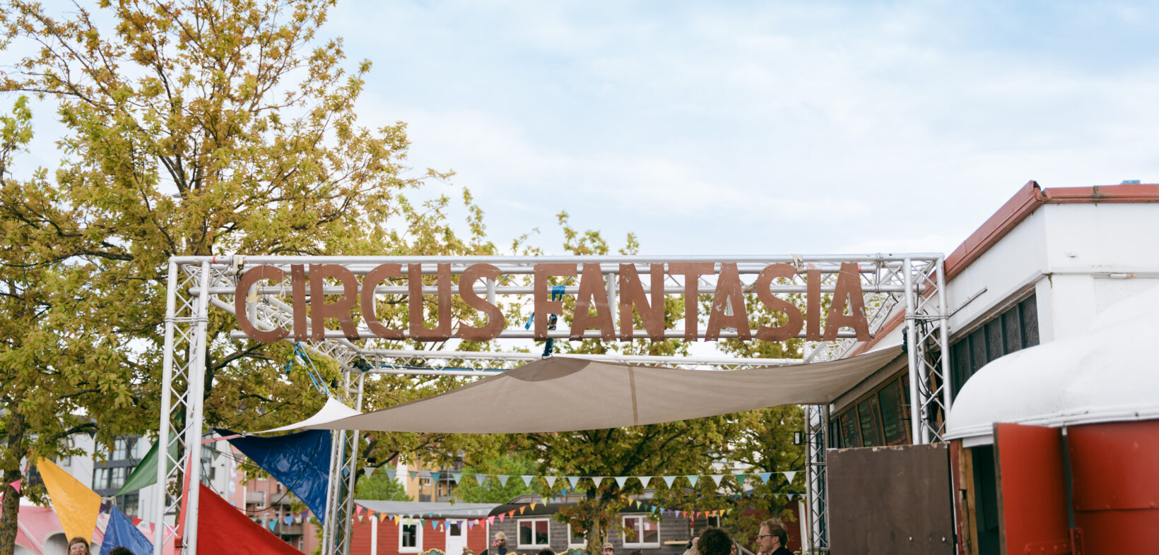 Der eiserne Schriftzug "Circus Fantasia" befestigt an einem Gerüst unter freiem Himmel - darunter Menschen, die den Circus-Platz erkunden