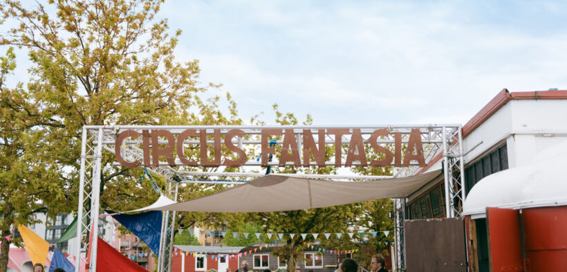 Der eiserne Schriftzug "Circus Fantasia" befestigt an einem Gerüst unter freiem Himmel - darunter Menschen, die den Circus-Platz erkunden