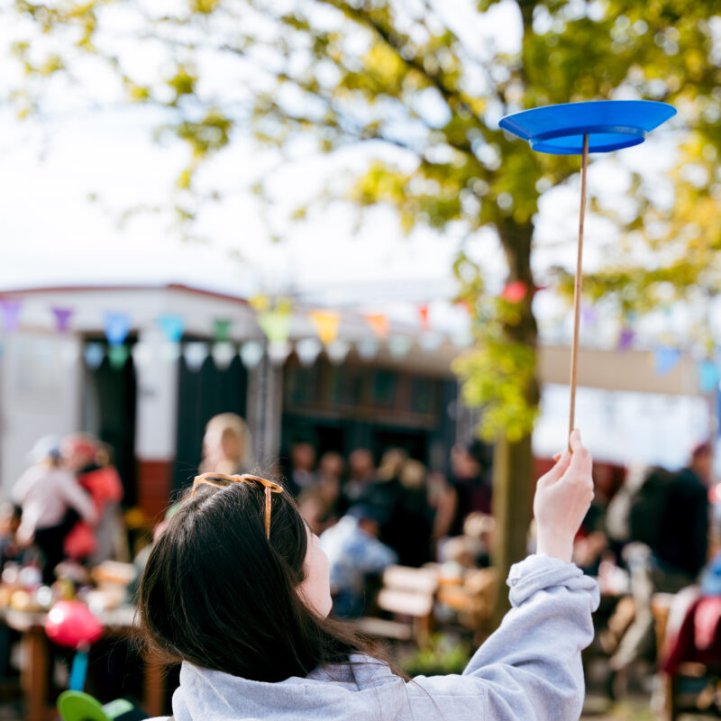Ein Mädchen ist von hinten zu sehen und dreht einen Teller auf einem Stab an, im Hintergrund ist der Circusplatz inklusive Menschen & bunten Wimpelketten zu sehen
