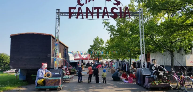 Ein großes, bogenförmiges Metallgerüst mit den metallenen Schriftzug "Circus Fantasia" steht unter freiem Himmel und bildet den Eingang zum Platz des Circus Fantasia, darunter stehen Menschen.